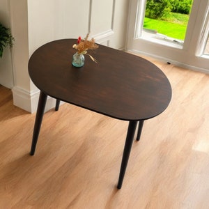May include: A dark brown oval coffee table with black legs. The table has a smooth surface and is set on a light wood floor. A small glass vase with dried flowers sits on the table. The table is near a window and a potted plant.