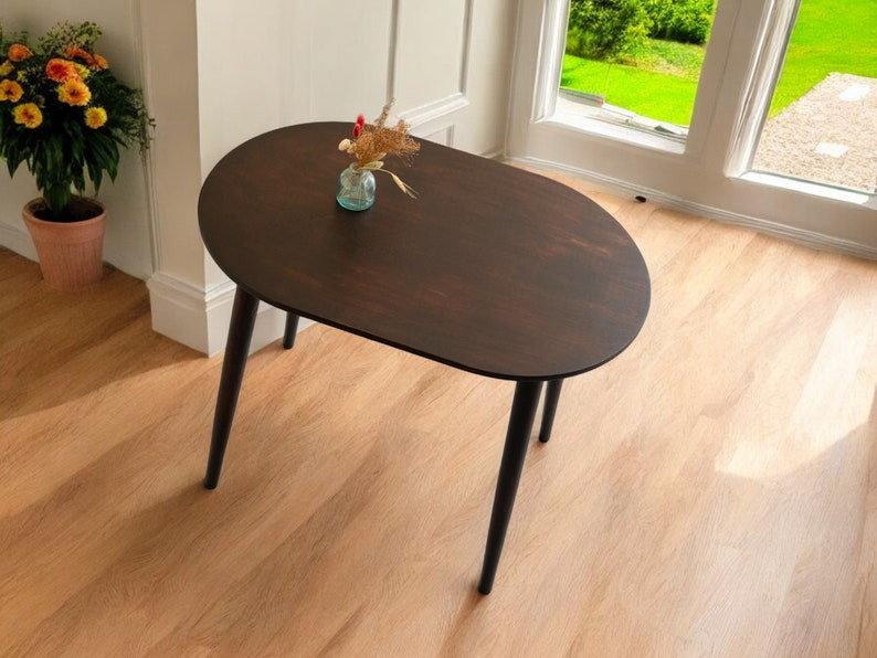 May include: A dark brown oval coffee table with black legs. The table has a smooth surface and is set on a light wood floor. A small glass vase with dried flowers sits on the table. The table is near a window and a potted plant.