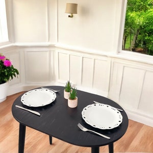 May include: A black oval dining table set for two with white plates, cutlery, and small potted succulents. The plates have a decorative edge. A pot of pink flowers sits in the corner. The room has white walls and a wooden floor.