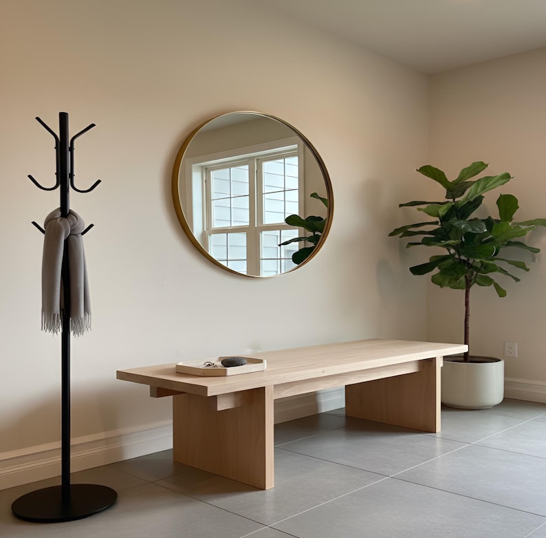 May include: A light wood bench with a rectangular top and two supporting legs. A round mirror with a gold frame hangs on the wall. A black coat rack holds a gray scarf. A potted plant is in the corner.