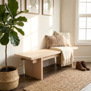 May include: A light wood bench with a cream-colored throw blanket and decorative pillows. A woven rug and a potted plant with large green leaves are in the foreground. Brown framed botanical prints hang on the wall.