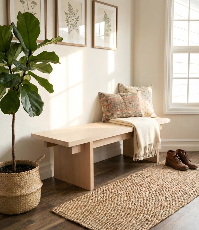 May include: A light wood bench with a cream-colored throw blanket and decorative pillows. A woven rug and a potted plant with large green leaves are in the foreground. Brown framed botanical prints hang on the wall.