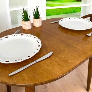 May include: An oval wooden dining table with two white plates featuring heart-shaped cutouts. Silver cutlery, including forks and knives, is laid out. Two small potted plants with green leaves are in the background.