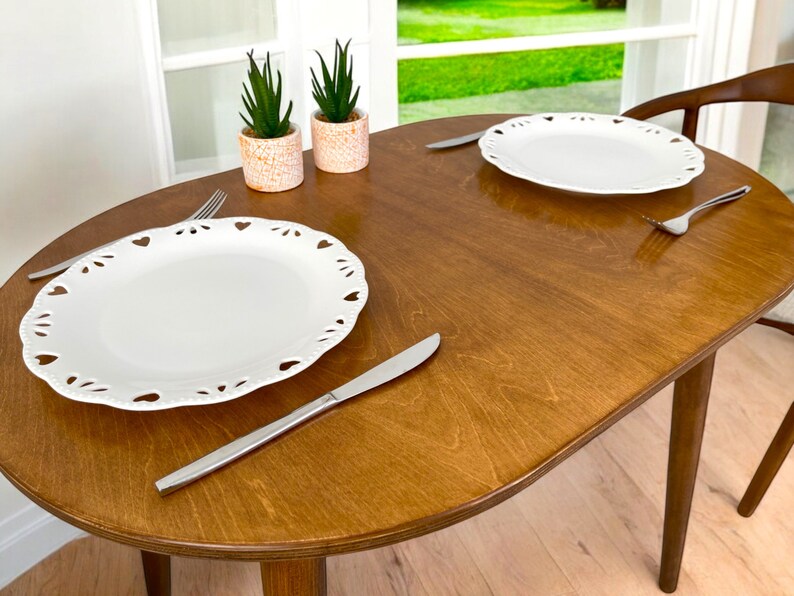 May include: An oval wooden dining table with two white plates featuring heart-shaped cutouts. Silver cutlery, including forks and knives, is laid out. Two small potted plants with green leaves are in the background.