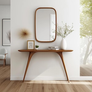 Peut inclure: Une entrée de style moderne du milieu du siècle avec une console en bois, un miroir rectangulaire et un vase blanc avec de la verdure. La table a un design incurvé et des pieds inclinés. Une photo encadrée et une petite plante sont sur la table.