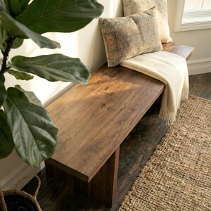 May include: A solid wood bench with a rich, dark brown finish. Two decorative pillows with patterned designs and a cream-colored throw blanket rest on the bench. A woven rug and a potted plant with large green leaves are in the foreground.