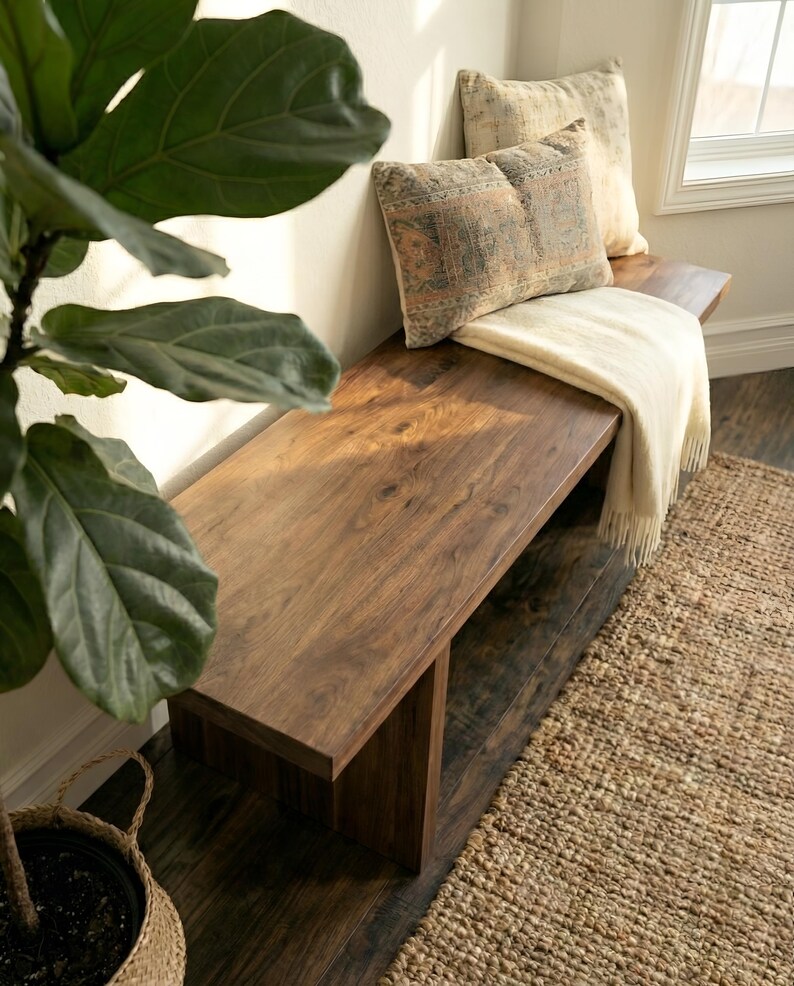May include: A solid wood bench with a rich, dark brown finish. Two decorative pillows with patterned designs and a cream-colored throw blanket rest on the bench. A woven rug and a potted plant with large green leaves are in the foreground.