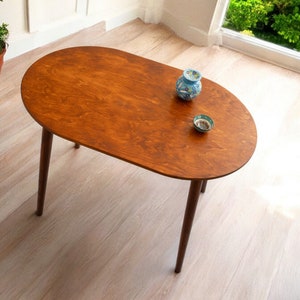 May include: Oval wooden coffee table with a rich brown finish and four slender, dark brown legs. The table has a smooth surface and is adorned with two small decorative ceramic pieces. The table is approximately 122 cm long.