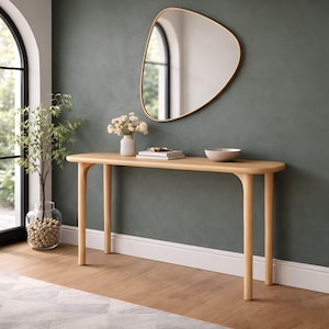May include: A light-toned wooden console table with rounded edges and four cylindrical legs. On the table surface sits a small white vase with flowers, a stack of books, and a shallow bowl. Above the table hangs an asymmetrical pebble-shaped mirror.