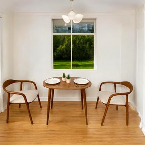 May include: A dining set featuring a small, round wooden table and two chairs. The chairs have curved wooden frames and cream-coloured upholstered seats. The table is set with plates and small potted plants. A window provides a view of green trees.