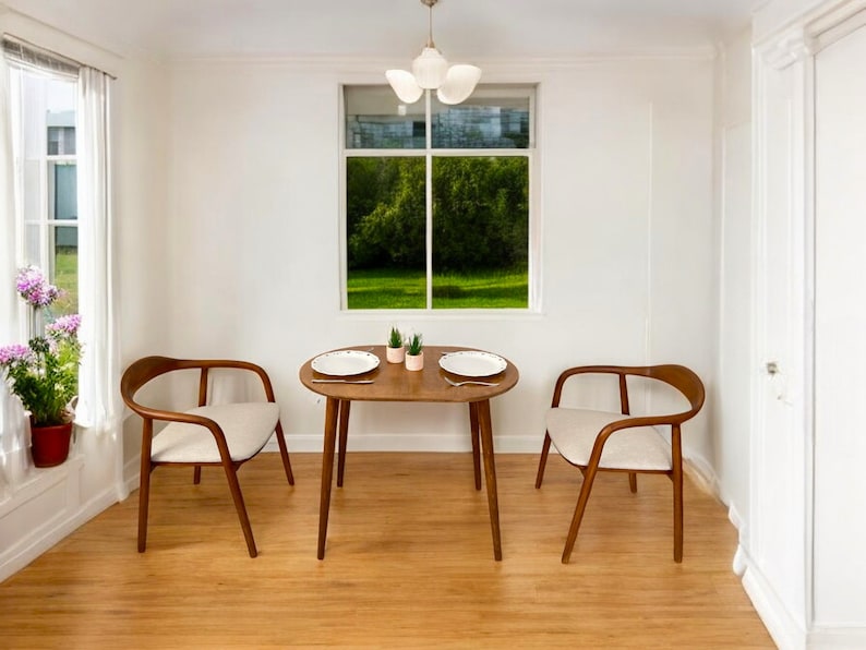 May include: A dining set featuring a small, round wooden table and two chairs. The chairs have curved wooden frames and cream-coloured upholstered seats. The table is set with plates and small potted plants. A window provides a view of green trees.