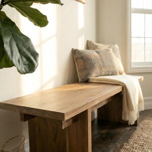 May include: A wooden bench with a light brown finish. Two decorative pillows with patterned fabric rest against the wall. A cream-colored throw blanket with fringe is draped over the bench. A large green plant is visible on the left.