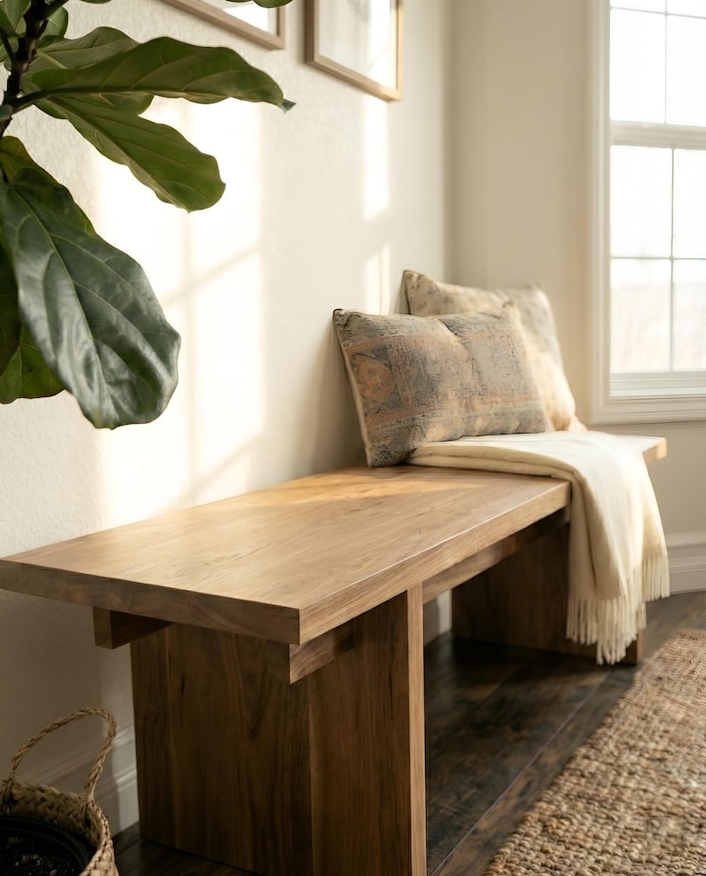 May include: A wooden bench with a light brown finish. Two decorative pillows with patterned fabric rest against the wall. A cream-colored throw blanket with fringe is draped over the bench. A large green plant is visible on the left.