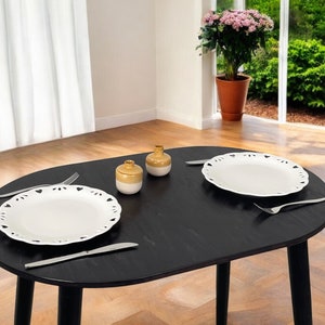 May include: A black oval dining table set for two. Two white plates with decorative edges are placed on the table, along with silverware and small ceramic jars. A potted plant and a window with a view are in the background.