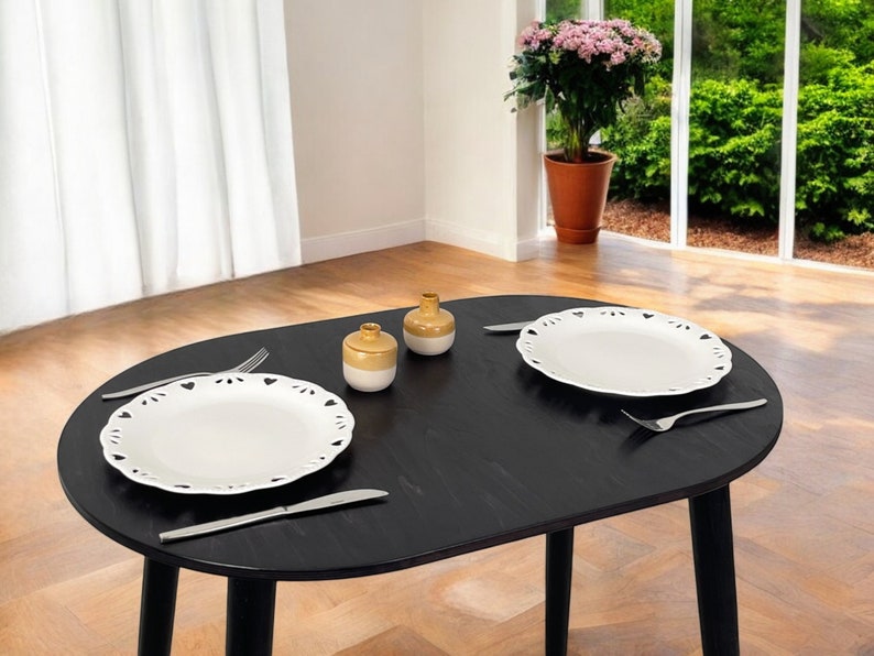 May include: A black oval dining table set for two. Two white plates with decorative edges are placed on the table, along with silverware and small ceramic jars. A potted plant and a window with a view are in the background.