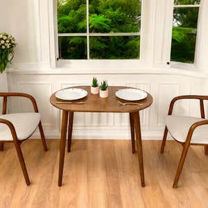 May include: A small dining set with a round wooden table and two chairs. The chairs have curved wooden frames and light-coloured upholstered seats. Two white plates and small potted plants are on the table. The room has white walls and a window.