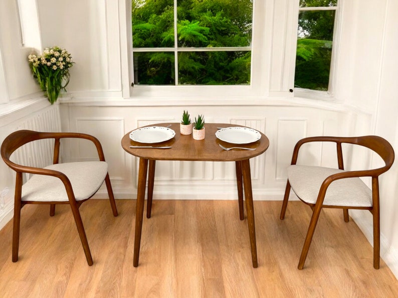 May include: A small dining set with a round wooden table and two chairs. The chairs have curved wooden frames and light-coloured upholstered seats. Two white plates and small potted plants are on the table. The room has white walls and a window.