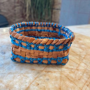 May include: A small, woven basket with a blue and brown pattern. The basket is made of natural materials and has a rectangular shape.