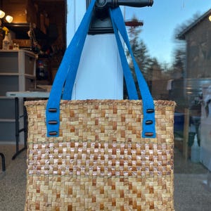 Pode incluir: Uma bolsa de mão de palha tecida com uma alça de tecido azul e botões de madeira castanho. A bolsa tem um padrão tecido com uma faixa de cor cobre.