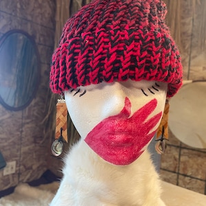 May include: A red and black knit beanie sits atop a white mannequin head. The mannequin has red lipstick and eye markings. Woven earrings with silver accents hang from the ears. The background is a rustic, wood-paneled wall.