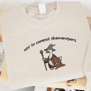 May include: Beige sweatshirt with the embroidered phrase "off to commit shenanigans" and a cartoon goose dressed as a wizard. Black and white sneakers are placed below the sweatshirt. The sweatshirt and sneakers are on a wooden shelf.