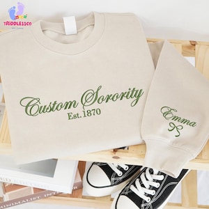 May include: A beige sweatshirt with the words "Custom Sorority Est. 1870" embroidered in green script. The sleeve displays the name "Emma" with a bow. Black and white sneakers are visible.