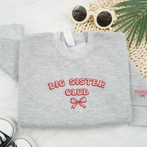 May include: A light gray sweatshirt with the words "BIG SISTER CLUB" embroidered in red, along with a red bow. The name "SOPHIA" is embroidered on the sleeve. The sweatshirt is surrounded by accessories, including sunglasses and baby shoes.