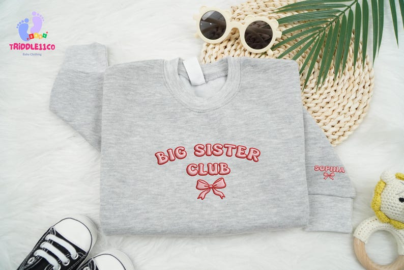 May include: A light gray sweatshirt with the words "BIG SISTER CLUB" embroidered in red, along with a red bow. The name "SOPHIA" is embroidered on the sleeve. The sweatshirt is surrounded by accessories, including sunglasses and baby shoes.