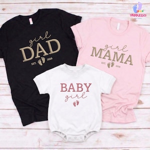 May include: A family set of matching shirts and a onesie. The black shirt says "girl DAD" with "EST. 2026" and footprints. The pink shirt says "girl MAMA" with "EST. 2026" and footprints. The white onesie says "BABY girl" with footprints.