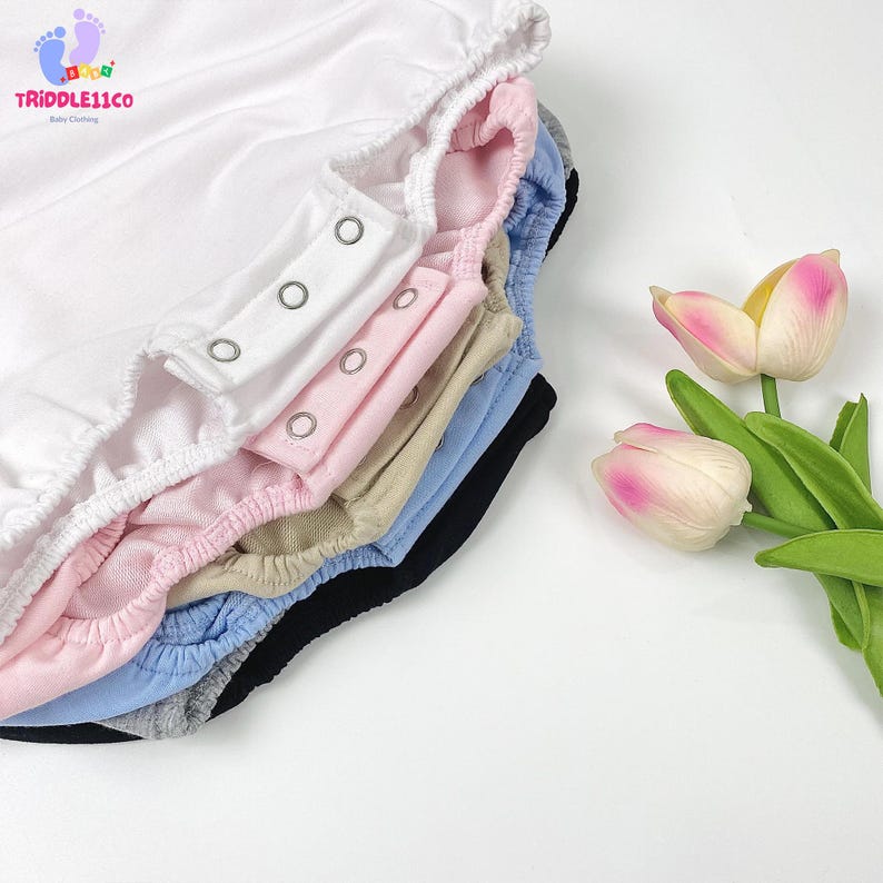 May include: A stack of baby bodysuits in white, light pink, beige, light blue, black, and grey, each featuring a row of silver metal snap fasteners at the crotch and elasticized leg openings, arranged next to two pink and yellow tulip flowers.