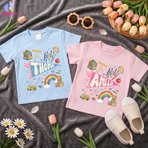 May include: A light blue t-shirt with the name Thomas and a pink t-shirt with the name Amelia, both featuring school-themed graphics including a backpack, crayons, a rainbow, and a school bus, alongside a pair of beige sunglasses and slip-on shoes.