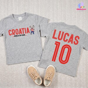 May include: Two heather gray short-sleeve t-shirts. The left shirt features the red text CROATIA, the text WORLD CUP 2026, and a dalmatian mascot graphic. The right shirt features the red text LUCAS and the number 10 in a bold, outlined font.