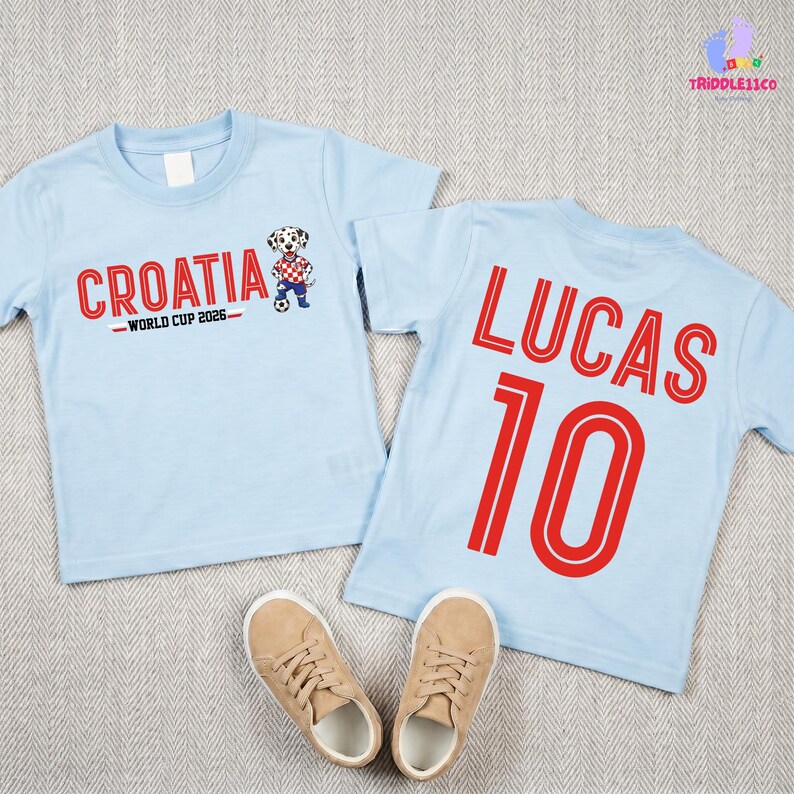 May include: Two light blue t-shirts. The front features the text CROATIA WORLD CUP 2026 with a dalmatian graphic. The back displays the name LUCAS and the number 10 in red. The shirts are paired with a pair of tan sneakers on a textured surface.