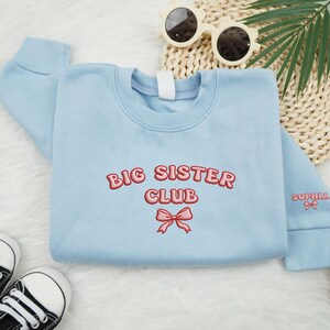 May include: A light blue sweatshirt with the words "BIG SISTER CLUB" embroidered in red. A pink bow is embroidered below the text. The sleeve has the name "Sophia" embroidered in red. Black and white sneakers and sunglasses are in the background.