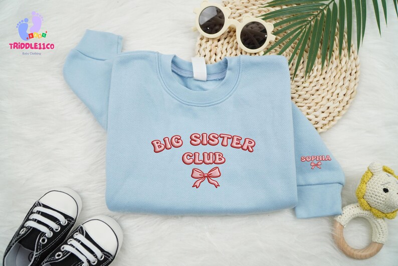 May include: A light blue sweatshirt with the words "BIG SISTER CLUB" embroidered in red. A pink bow is embroidered below the text. The sleeve has the name "Sophia" embroidered in red. Black and white sneakers and sunglasses are in the background.