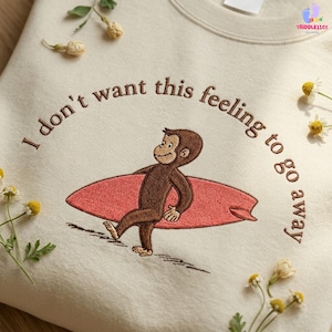 May include: A cream-colored sweatshirt featuring brown embroidery of a monkey carrying a pink surfboard. The text "I don't want this feeling to go away" is embroidered in a brown arc above the monkey. Small white flowers are scattered around.
