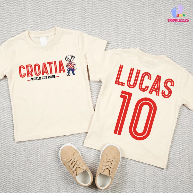 May include: Two cream-colored short-sleeve t-shirts. One shirt features the red text "CROATIA WORLD CUP 2026" with a cartoon dalmatian soccer player. The other shirt displays the red name "LUCAS" above the red number "10".
