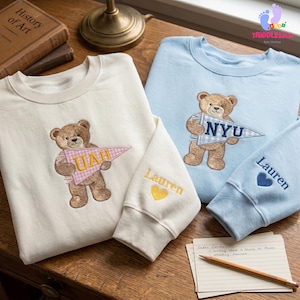 May include: Cream and light blue sweatshirts with embroidered teddy bears holding pennants. One sweatshirt has "UAH" and the other "NYU". Sleeves feature names and heart details. The sweatshirts are on a wooden surface.
