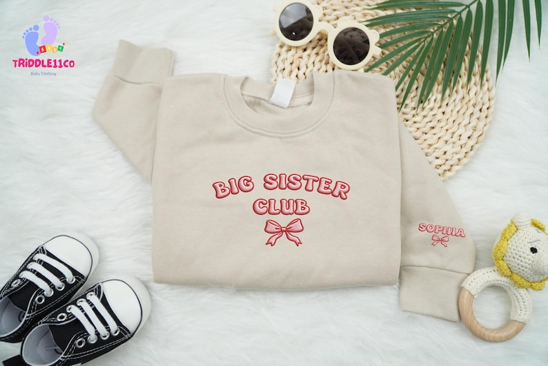 May include: Beige sweatshirt with the text "BIG SISTER CLUB" in red embroidery, featuring a bow graphic. The sleeve is embroidered with the name "SOPHIA" and a bow. Includes black and white sneakers, sunglasses, and a baby toy.