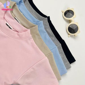 May include: A set of five short-sleeved t-shirts in pink, beige, light blue, gray, and black, each featuring a ribbed crew neckline. Beside the shirts, a pair of cream-colored sunglasses with round frames and bear-ear accents is displayed.