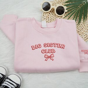 May include: A light pink sweatshirt with the words "BIG SISTER CLUB" embroidered in red. A red bow is embroidered below the text. The name "Sophia" is embroidered on the sleeve. Black and white sneakers and sunglasses are also in the image.