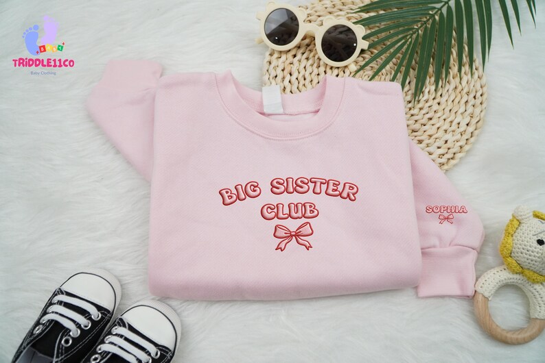 May include: A light pink sweatshirt with the words "BIG SISTER CLUB" embroidered in red. A red bow is embroidered below the text. The name "Sophia" is embroidered on the sleeve. Black and white sneakers and sunglasses are also in the image.