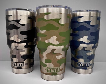 Puede incluir: Tres vasos YETI con diferentes patrones de camuflaje. Los vasos son de acero inoxidable y tienen un logotipo YETI negro en la parte inferior.