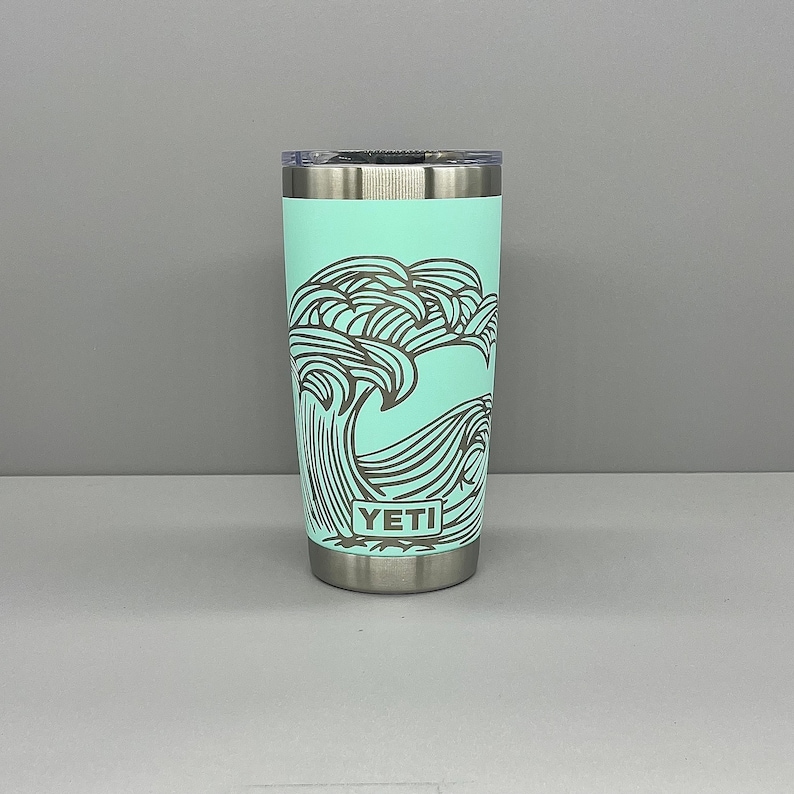 K&ouml;nnte beinhalten: Ein t&uuml;rkisfarbener YETI-Becher mit einem schwarz-wei&szlig;en Wellendesign. Der Becher hat einen Deckel und einen Boden aus Edelstahl.