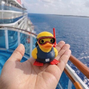 Puede incluir: Una peque&ntilde;a figura de pato de goma amarillo vestido de buceador. El pato lleva un chaleco azul oscuro, aletas rojas y una m&aacute;scara de buceo roja y negra con un tubo de snorkel rojo a juego sujeto al lado de la cabeza.