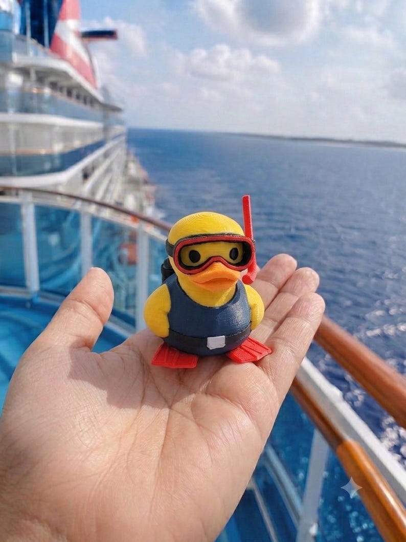 Puede incluir: Una peque&ntilde;a figura de pato de goma amarillo vestido de buceador. El pato lleva un chaleco azul oscuro, aletas rojas y una m&aacute;scara de buceo roja y negra con un tubo de snorkel rojo a juego sujeto al lado de la cabeza.
