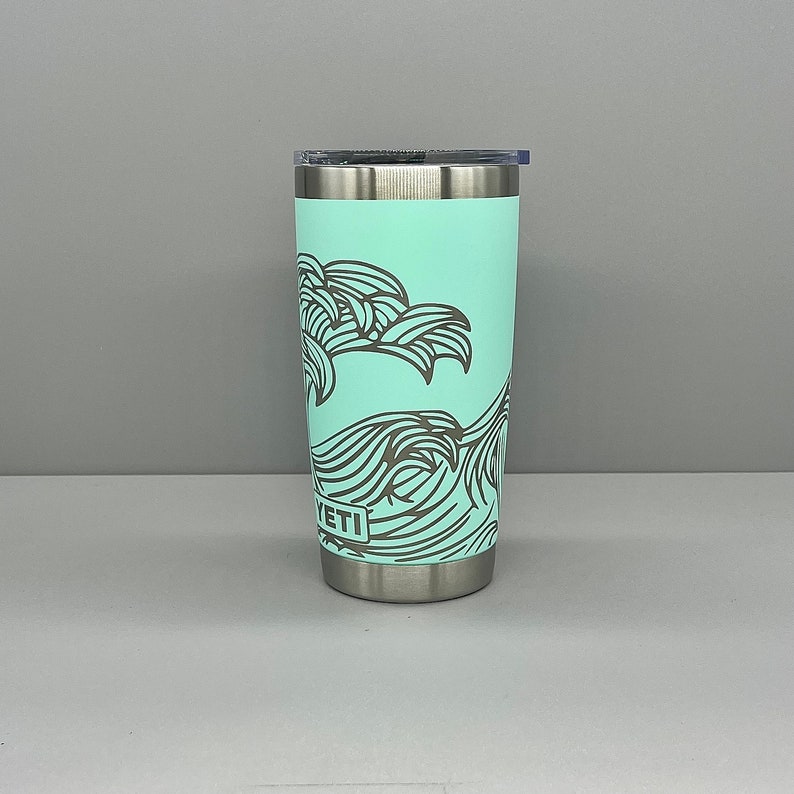K&ouml;nnte beinhalten: Ein Edelstahlbecher mit hellblauer Au&szlig;enseite und einem schwarz-wei&szlig;en Wellendesign. Der Becher hat einen Deckel und das Wort "YETI" ist auf der Seite aufgedruckt.