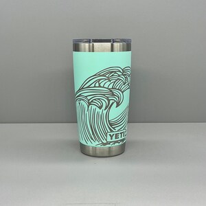 K&ouml;nnte beinhalten: Ein t&uuml;rkisfarbener Yeti Rambler Becher mit einem schwarz-wei&szlig;en Wellendesign und dem Wort "YETI" auf der Seite.