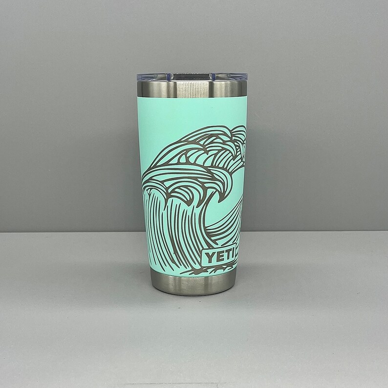 K&ouml;nnte beinhalten: Ein t&uuml;rkisfarbener Yeti Rambler Becher mit einem schwarz-wei&szlig;en Wellendesign und dem Wort "YETI" auf der Seite.