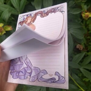 May include: A purple and white notepad with a decorative border featuring a woman with long brown hair and a purple dress. The notepad has lined pages and a gold Triforce symbol.